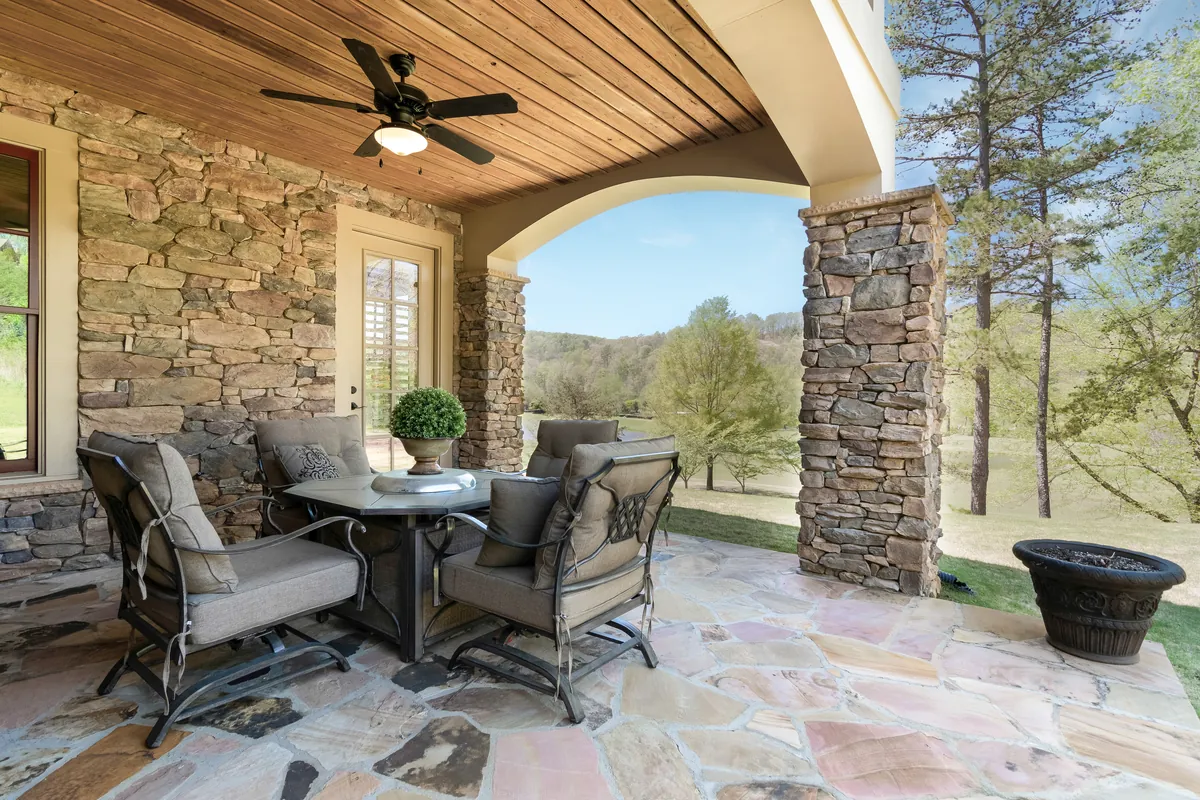 Covered stone patio with a dining set overlooking trees, lawn, and a pond