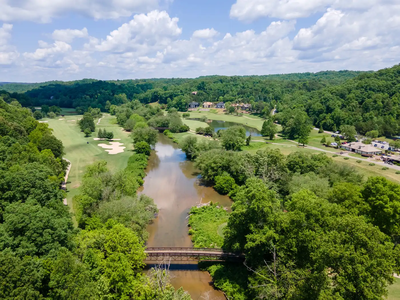 Aerial view of a wide river winding between wooded banks past golf fairways with sand bunkers, crossed by a metal footbridge, with homes on a rise beyond