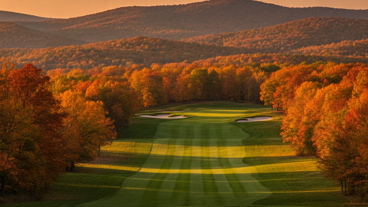 A golf fairway with mown stripes and pale sand bunkers running toward a distant green, surrounded by peak-autumn orange and red hardwoods with layered mountain ridges behind at sunset